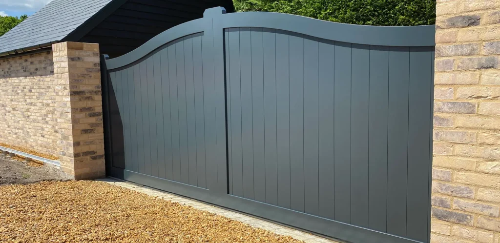 A grey aluminium gate with an elegant curved top, installed between brick pillars for a stylish entrance. Gates Wellington offers high-quality aluminium gates.